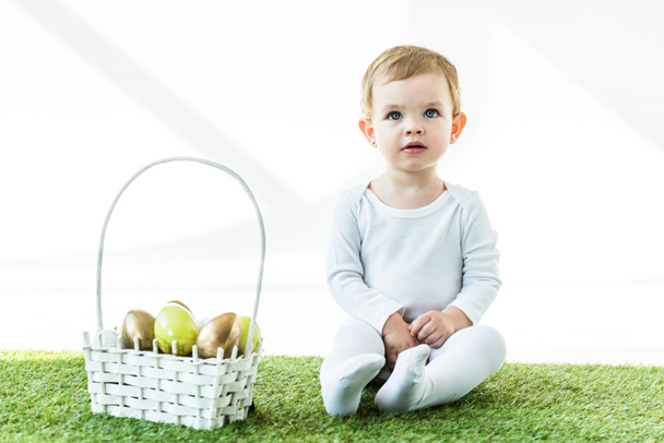 cute child sitting near straw basket with bright Easter eggs isolated on white - Photo, Image