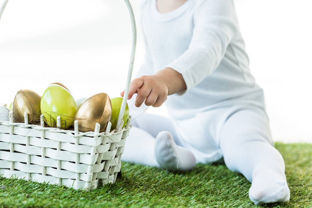 cropped view of kid sitting near straw basket with yellow and golden chicken eggs isolated on white - Photo, Image