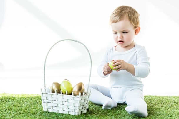 adorable bebé sosteniendo huevo de pollo amarillo mientras está sentado cerca de la canasta de paja con huevos de Pascua aislados en blanco
 - Foto, Imagen