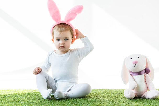 adorable niño en conejito orejas diadema sentado cerca divertido juguete conejo aislado en blanco
 - Foto, Imagen