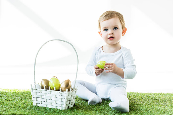 adorable kid holding yellow chicken egg while sitting on green grass near straw basket with Easter eggs isolated on white - Fotó, kép