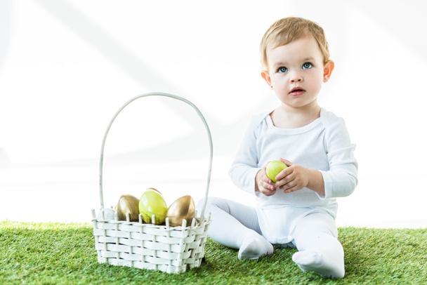 lindo bebé rubio sosteniendo huevo de pollo amarillo mientras está sentado cerca de la canasta de paja con huevos de Pascua aislados en blanco
 - Foto, Imagen