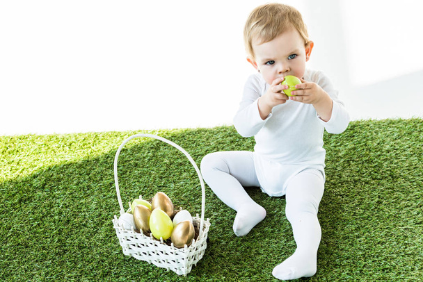 cute child holding yellow kitchen egg near face while sitting near straw basket with Easter eggs isolated on white - Foto, afbeelding