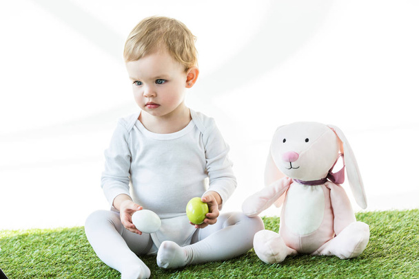 мила дитина тримає курячі яйця, сидячи на зеленій траві біля іграшкового кролика ізольовано на білому
 - Фото, зображення