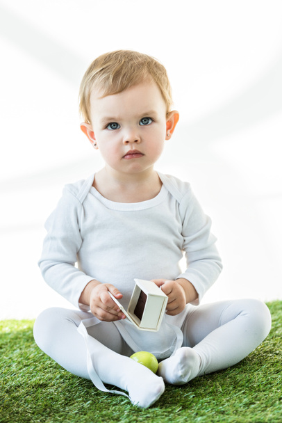 adorable kid sitting on green grass with white cardboard box and yellow chicken egg isolated on white - Foto, Imagem