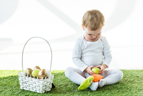 прелестный ребенок сидит рядом с соломенной корзиной с красочными пасхальными яйцами, изолированными на белом
 - Фото, изображение