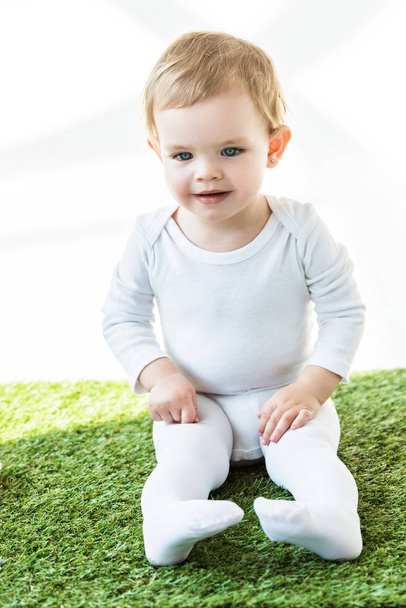 adorable smiling baby with blonde hair sitting on green grass isolated on white - Foto, afbeelding