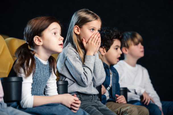 Niedliches Kind, das die Hände vor dem Gesicht hält, während es sich mit multikulturellen Freunden einen Film ansieht - Foto, Bild