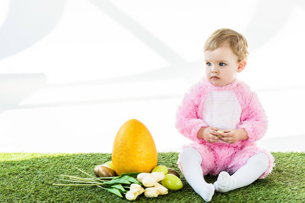 aranyos baba rózsaszín bolyhos jelmez ül közel sárga strucc tojás, színes csirke tojás és a tulipán elszigetelt fehér - Fotó, kép