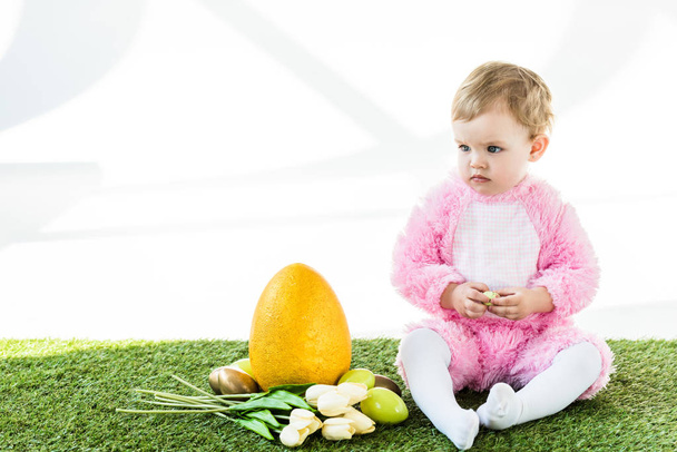 adorable child in pink fluffy costume sitting near yellow ostrich egg, colorful chicken eggs and tulips isolated on white - Foto, afbeelding