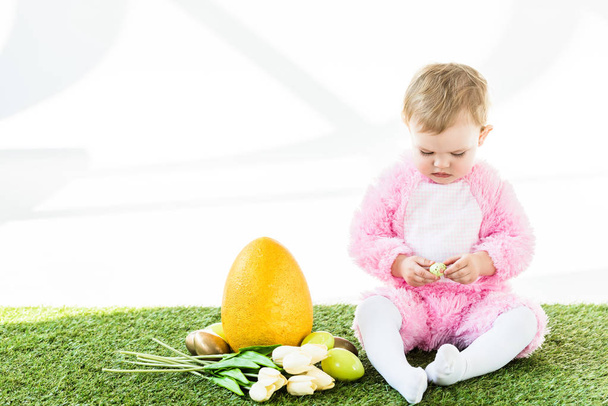 imádnivaló gyermek rózsaszín bolyhos jelmez ül közel sárga strucc tojás, színes csirke tojás és a tulipán elszigetelt fehér - Fotó, kép