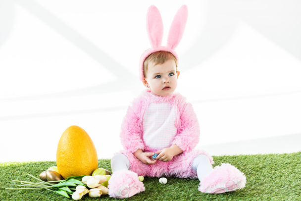 adorable niño en divertido traje de conejo sentado cerca de coloridos huevos de pollo, tulipanes y huevo de avestruz amarillo aislado en blanco
 - Foto, Imagen
