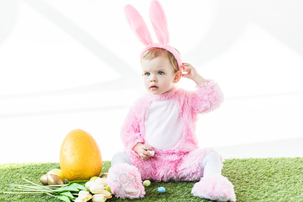 lindo bebé en divertido traje de conejo sentado cerca de los huevos de Pascua, tulipanes y huevo de avestruz amarillo aislado en blanco
 - Foto, Imagen