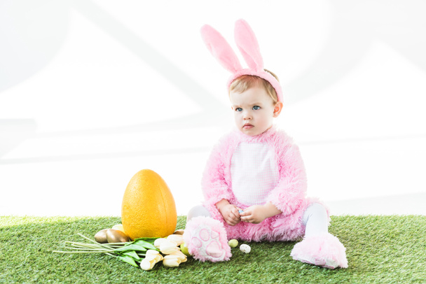 lindo bebé en divertido traje de conejo sentado cerca de huevo de avestruz amarillo, coloridos huevos de Pascua y tulipanes aislados en blanco
 - Foto, Imagen