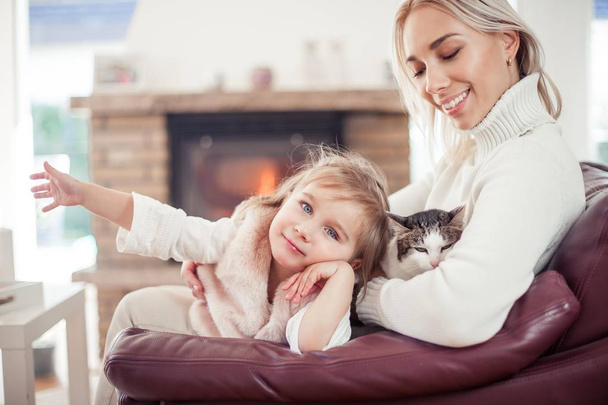 Schöne Mutter, kleine Tochter und eine Katze sitzen auf dem Sofa neben dem Kamin. Familie im häuslichen Bereich. gemütlich. - Foto, Bild