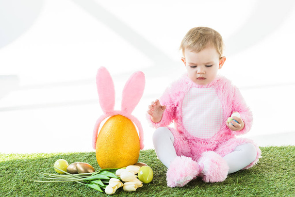 aranyos gyerek rózsaszín bolyhos jelmez ült a közelben sárga strucc tojás nyuszi füle fejpánt, színes húsvéti tojást és a tulipán elszigetelt fehér - Fotó, kép