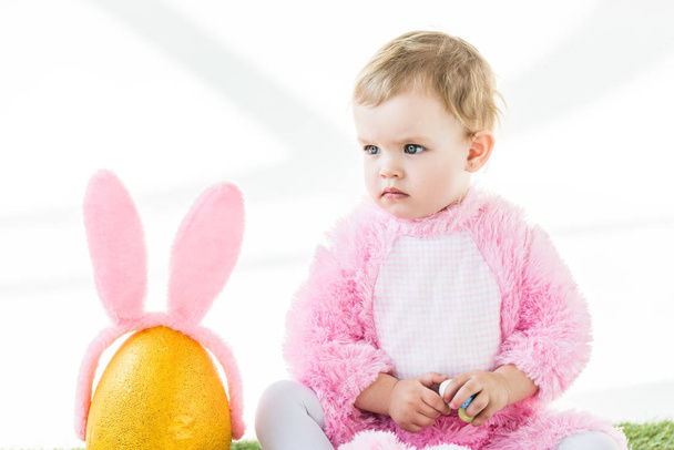 niedliches Kind im rosafarbenen flauschigen Kostüm, das bunte Wachteleier hält, während es in der Nähe eines gelben Straußeneiers mit Hasenohren-Stirnband sitzt, das auf weiß isoliert ist - Foto, Bild