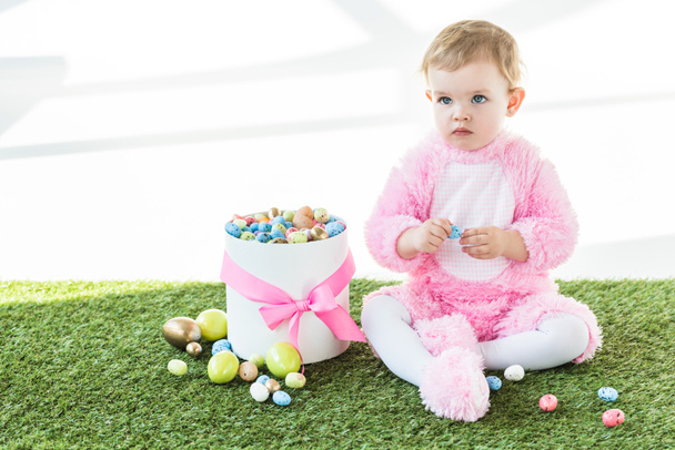 imádnivaló baba rózsaszín bolyhos jelmezben ül a zöld fű közelében doboz színes húsvéti tojást izolált fehér - Fotó, kép