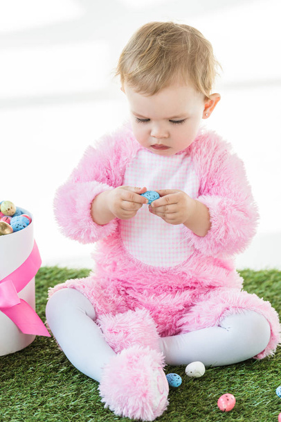 cute kid in pink fluffy costume holding blue quail egg while sitting near box with Easter eggs isolated on white - Foto, immagini