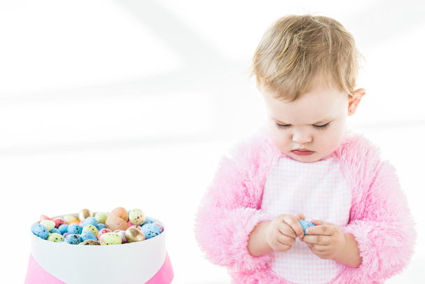 adorable niño en traje mullido rosa sosteniendo huevo de codorniz azul mientras está sentado cerca de la caja con coloridos huevos de Pascua aislados en blanco
 - Foto, Imagen