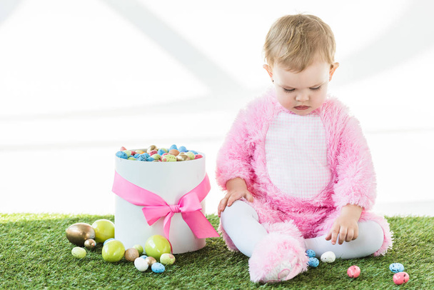 aranyos baba rózsaszín bolyhos jelmezben ül a doboz közelében színes húsvéti tojást elszigetelt fehér - Fotó, kép