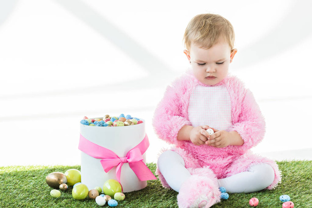 lindo bebé en traje mullido rosa sosteniendo huevo de codorniz mientras está sentado cerca de la caja con coloridos huevos de Pascua aislados en blanco
 - Foto, Imagen