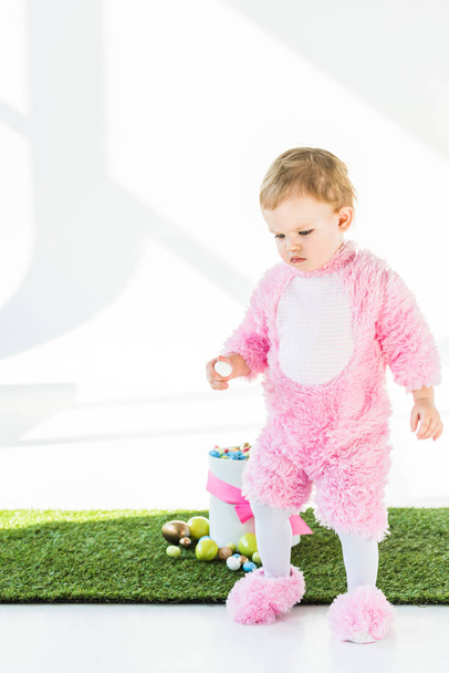 adorable niño en traje mullido rosa sosteniendo huevo de codorniz mientras está de pie cerca de la caja con coloridos huevos de Pascua aislados en blanco
 - Foto, Imagen