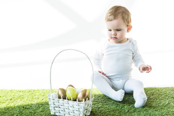 adorable child sitting on green grass near straw basket with yellow and golden Easter eggs isolated on white  - Fotó, kép
