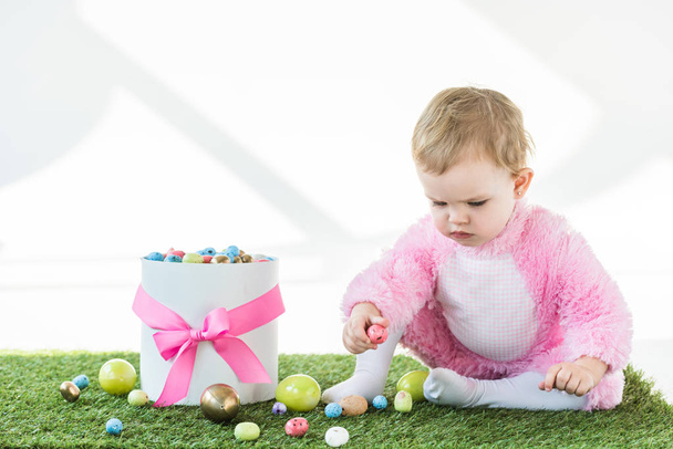 cute baby in pink fluffy costume sitting on green grass near gift box with pink bow and colorful Easter eggs isolated on white - Photo, Image