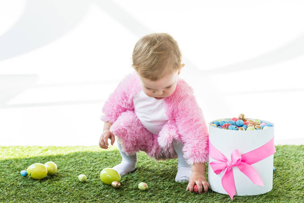 imádnivaló gyerek, rózsaszín bolyhos jelmezben figyelembe színes tojásokat a zöld fű közelében ajándékdoboz rózsaszín orr elszigetelt fehér - Fotó, kép