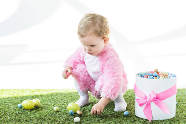 adorable niño en traje mullido rosa tomando huevos de colores de hierba verde cerca de caja de regalo con arco rosa aislado en blanco
 - Foto, Imagen
