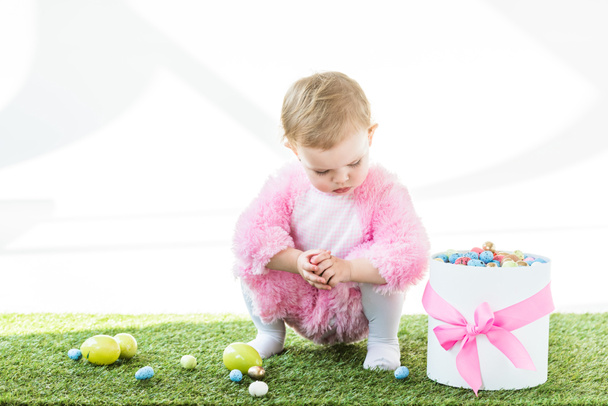 aranyos baba rózsaszín bolyhos jelmezben ülve a zöld fű közelében ajándékdoboz rózsaszín íj és színes húsvéti tojást elszigetelt fehér - Fotó, kép