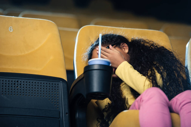 scared african american child holding hand on face while sitting in cinema seat with paper cup in cup holder - Zdjęcie, obraz