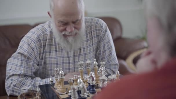 Two old men playing chess sitting at home on the leather sofa. Bearded man thinking which chess piece to make a move. Caucasian old men neighbors friends playing chess joyfully indoors - Video