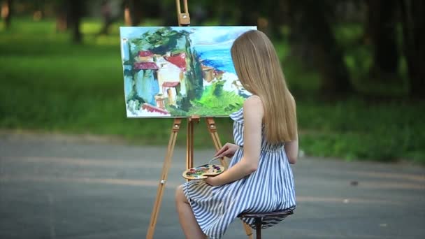 Prachtig blond haar meisje zit op kruk en tekent u een afbeelding in het park met behulp van een palet met verf en een spatel - Video