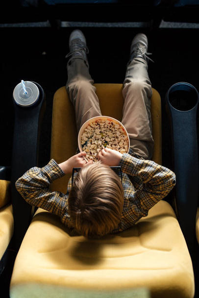 overhead view of boy eating popcorn while sitting in cinema chair near paper cup - Photo, Image