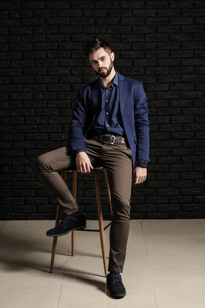 Fashionable young man sitting on chair against dark brick wall - Zdjęcie, obraz