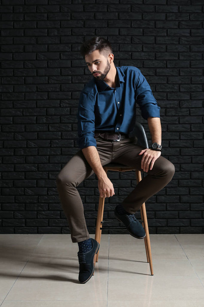 Divatos fiatalember ül a szék sötét téglafal ellen - Fotó, kép