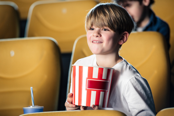 adorable smiling boy holding stripped paper cup while watching movie in cinema - Фото, зображення