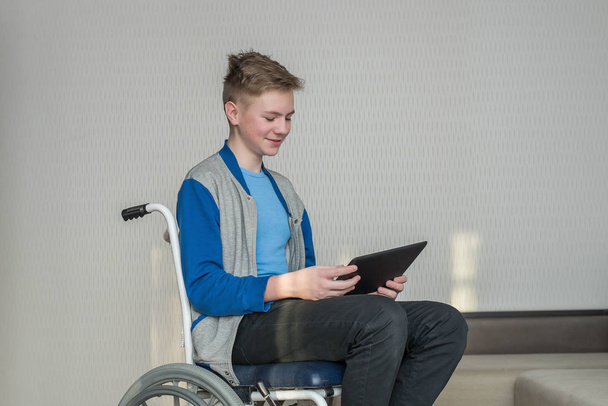 teini poika opiskelija istuu pyörätuolissa, kun käytät tablettia kotona. - Valokuva, kuva