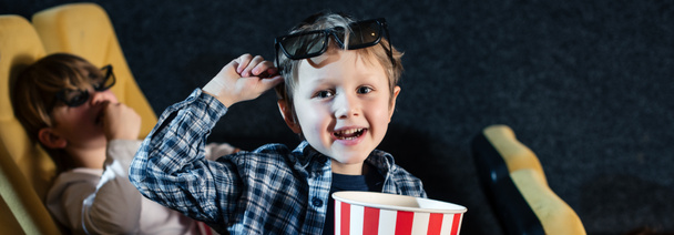 panoramic shot of cute boy looking at camera and holding stripped paper cup with pop corn - Foto, imagen