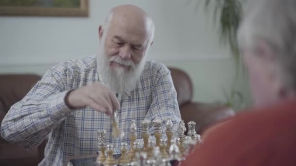 Adult friends playing chess sitting at home on the leather sofa. Bearded man thinking which chess piece to make a move. - Кадры, видео