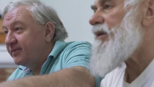 Close up of two mature senior men watching TV and discussing at home. Friends watching football soccer while eating chips. People involved in game - Кадри, відео