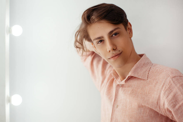 Portrait of masculine attractive european young man with brown eyes, dreaming face expression, smiling. Leaning on a wall, against blank background with light bulb in striped shirt indoor. People - Foto, immagini