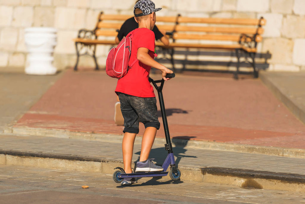 A guy in denim shorts and a red t-shirt rides a scooter. Extreme sport . The scooter is in motion. Boys against a wooden bench. - Photo, Image