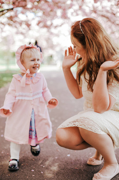 Loving mother and baby girl on background blooming pink flowers of cherry tree at springtime. Beautiful woman with daughter among garden flowers outdoors. Family on nature in Greenwich park, UK - Zdjęcie, obraz