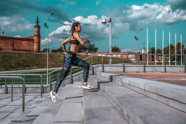 Athletic girl, runs in the city in the summer, morning afternoon jogging. Against the sky on the stairs. Active and healthy lifestyle. Clothing leggings top, sneakers. In headphones to the music. - Fotografie, Obrázek