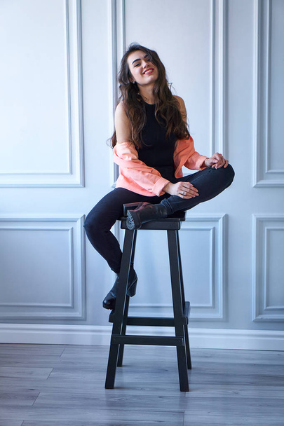 Portrait of attractive smiling brunette girl in peach jacket sitting on chair - Fotó, kép