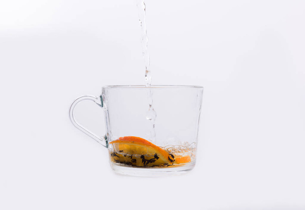 Agua que se vierte en una taza de vidrio con té y rodajas de naranja aisladas en blanco
 - Foto, Imagen