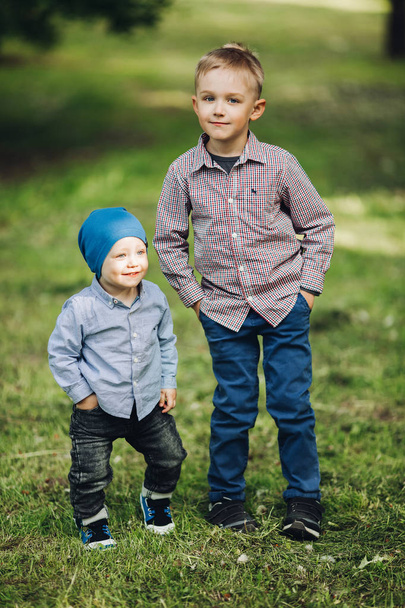 zwei kleine Jungen in lässigem Look posieren im Park und halten die Hände in den Taschen. - Foto, Bild